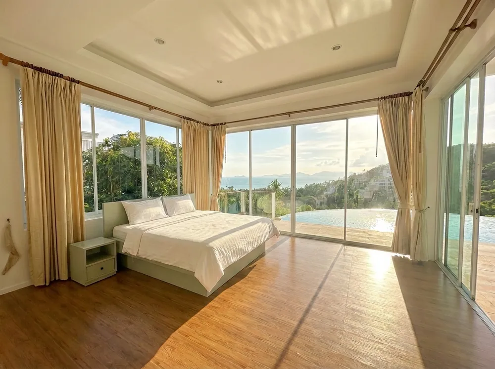 Bright bedroom with large windows, beige curtains, wooden floor, bed, and ocean view with pool outside.