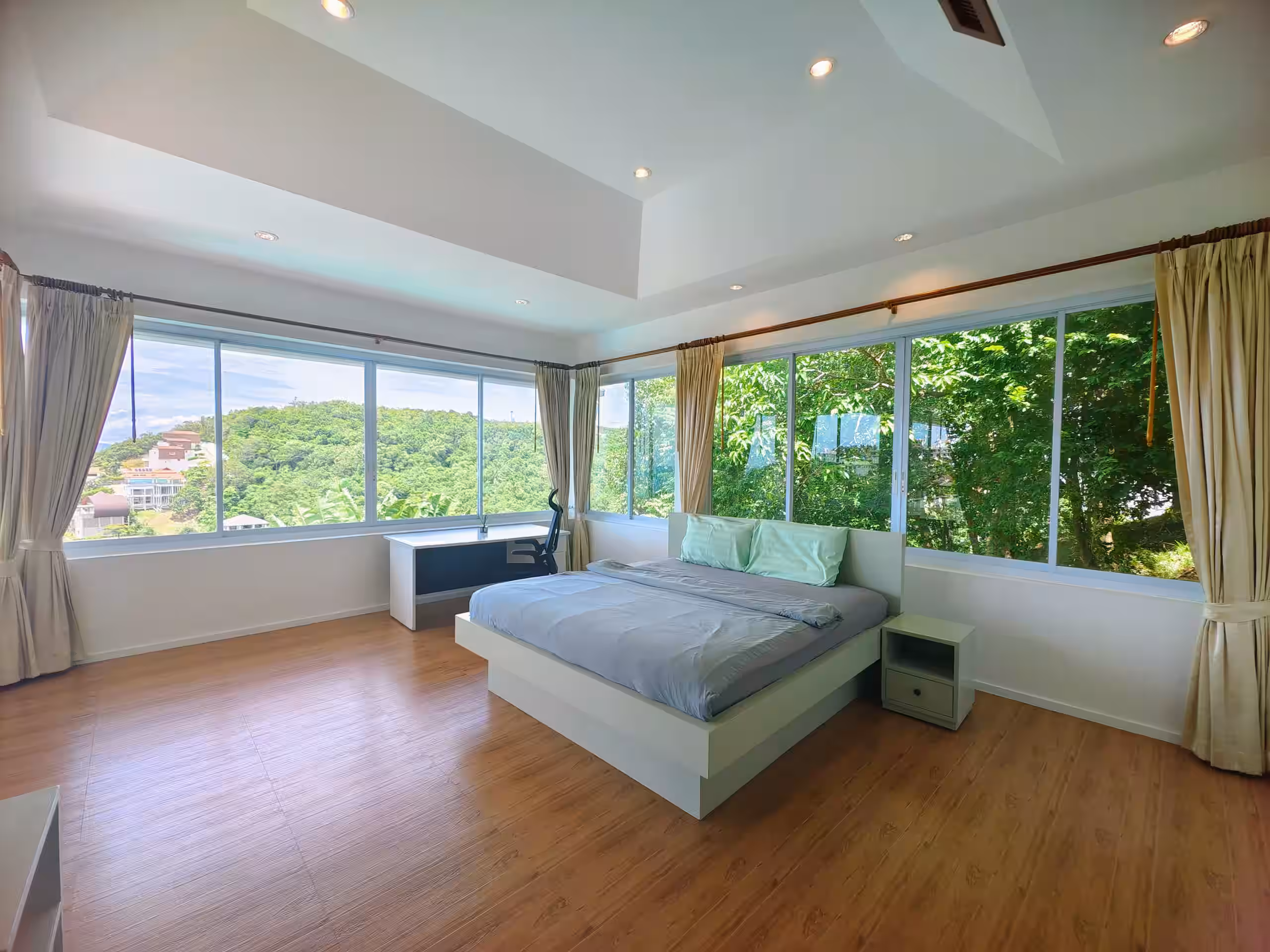 Bright bedroom with large windows, gray bed, light green pillows, wooden floor, desk, and chair.