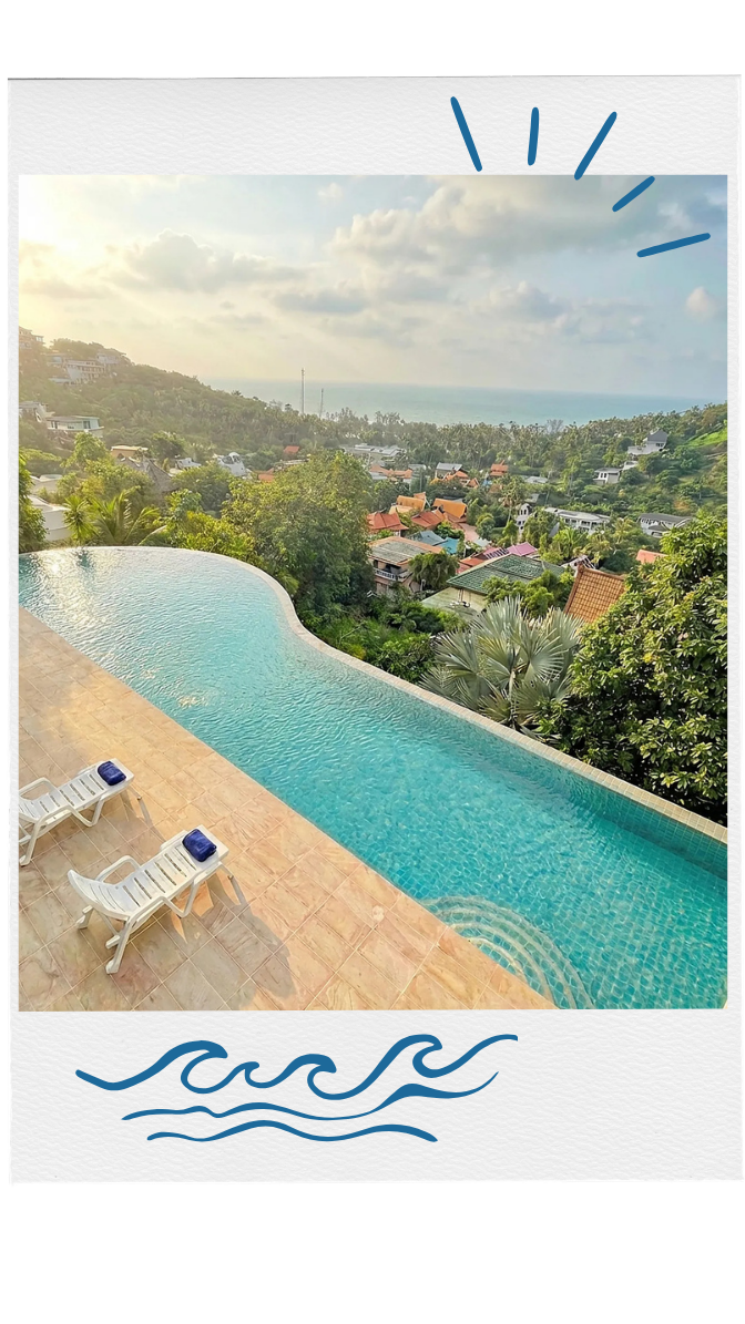 Infinity pool overlooking green hills, houses, and distant ocean under a cloudy sky.