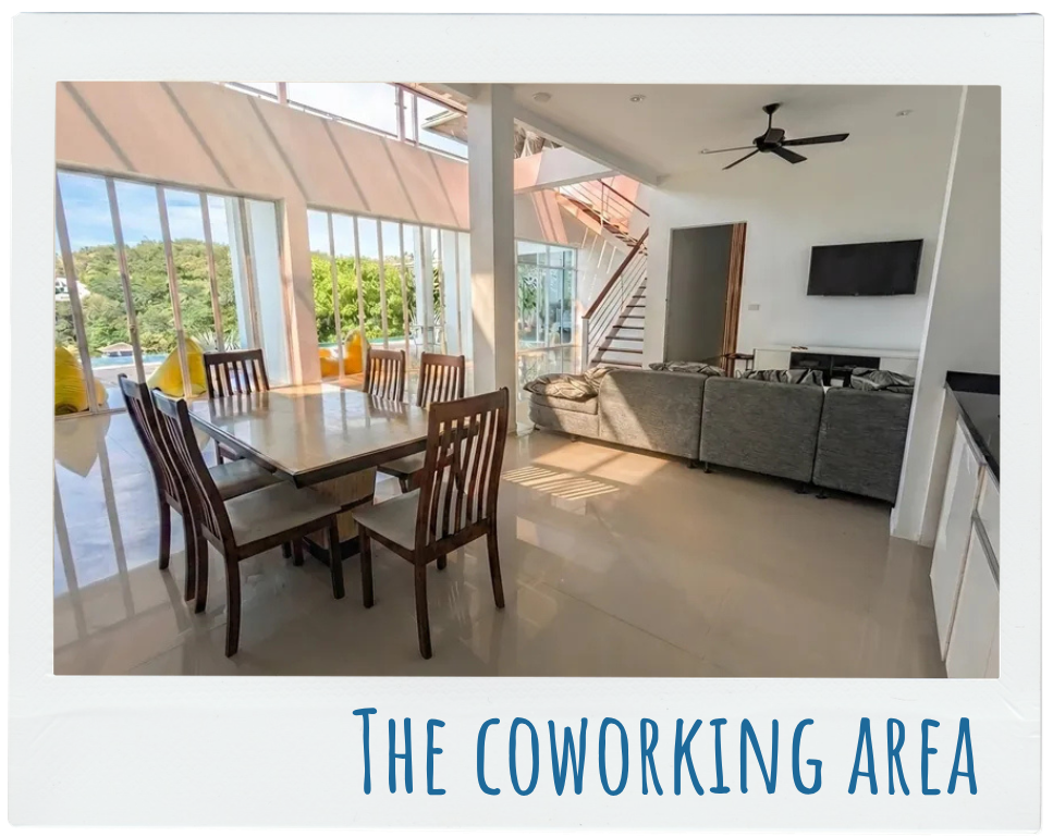 Bright coworking area with wooden table and chairs by large windows and a gray sofa facing a wall TV.